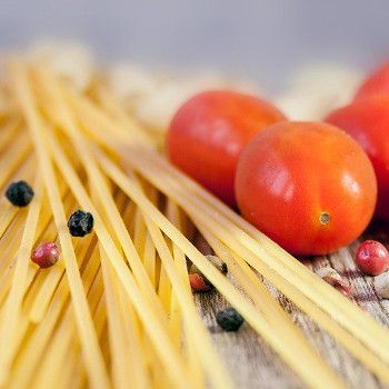 Na drewnianym stole leży spaghetti, kilka pomidorków koktajlowych i ziarenka czerwonego i czarnego pieprzu.