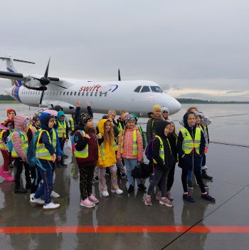 Uczniowie stoją na płycie lotniska przy samolocie.