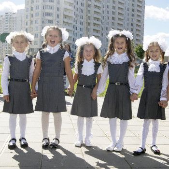 Dziewczynki w odświętnych ubraniach i z białymi kokardami we włosach, stoją przed wysokim budynkiem. Są uśmiechnięte, trzymają się za ręce i wyglądają jakby nie mogły doczekać się pójścia do szkoły.